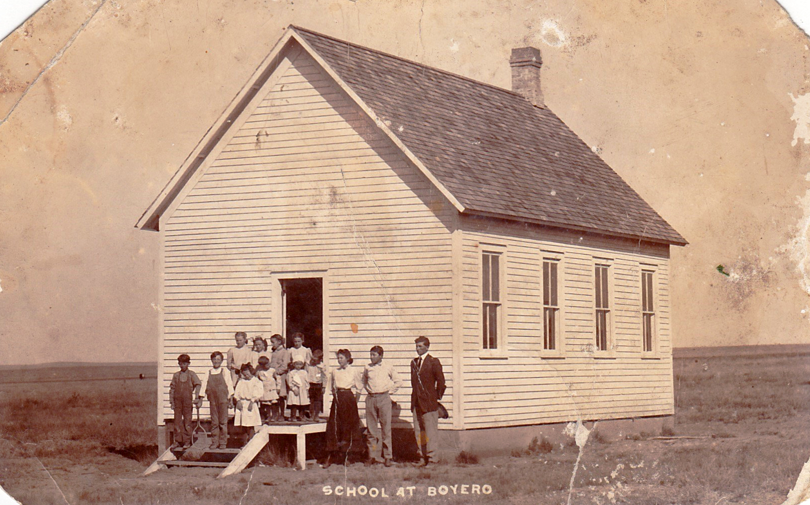 Historic photo of students and teachers standing outside the one-room schoolhouse in Boyero, Colorado, early 1900s, surrounded by open prairie landscape.